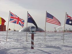 The ceremonial South Pole.  Flags of the Antarctic Treaty signatories are arrayed around it, and the Pole Station's old dome is in the background.
